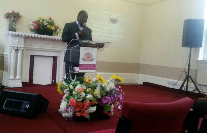 A minister speaking from the platform beside the flower arrangements