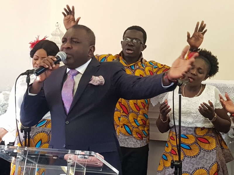 A pastor preaching with his arm raised as the choir worships behind him at Divine Encounter